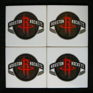 May include: Four square white coasters featuring the Houston Rockets logo. The logo includes a red stylized letter "R" over a dark basketball, with "HOUSTON ROCKETS" in white text on a black banner.