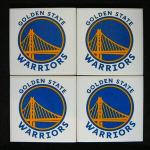 May include: Four square coasters featuring the Golden State Warriors logo. Each coaster has a white background with the team's logo: a blue circle with a yellow bridge and the words "Golden State Warriors" in blue.