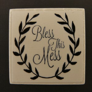 May include: White square tile with black lettering that reads "Bless This Mess" surrounded by a black wreath.