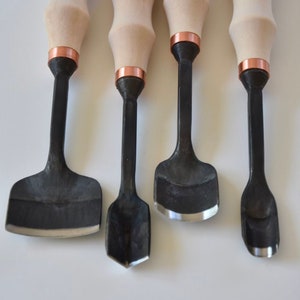 Może przedstawiać: Zestaw czterech narzędzi do rzeźbienia z drewnianymi uchwytami i czarnymi metalowymi ostrzami. Narzędzia mają miedziane pasmo na górze uchwytu. Narzędzia są oznaczone literą "E" i słowami "Made in Lithuania" i "Pagaminta Lietuvoje".