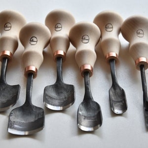 May include: Set of seven wood carving tools with black metal blades and wooden handles. The handles are a light brown color and have a copper band around the top. The blades are all different shapes and sizes.