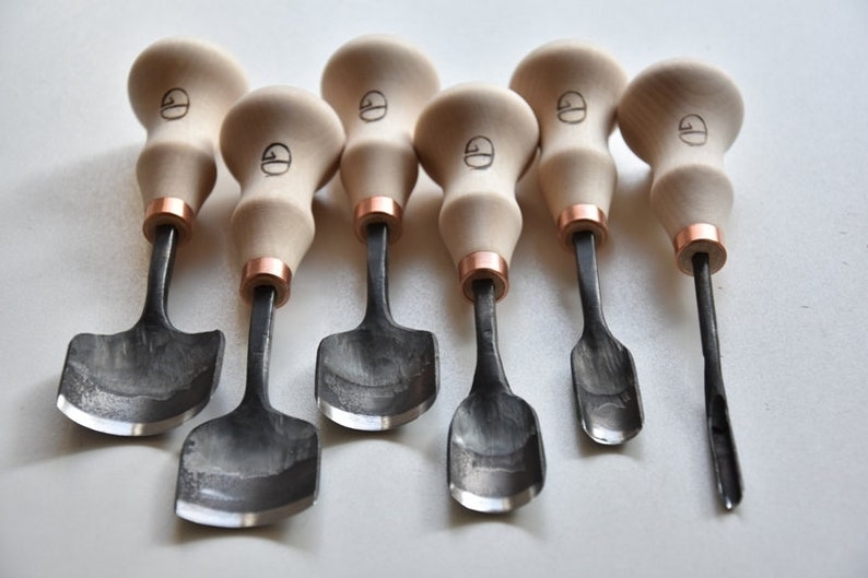 May include: Set of seven wood carving tools with black metal blades and wooden handles. The handles are a light brown color and have a copper band around the top. The blades are all different shapes and sizes.