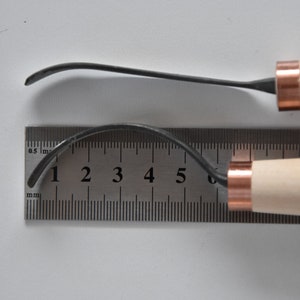 May include: Two wood carving tools with copper accents. The tools are laid out on a ruler with markings in millimetres. The top tool is 6.5 cm long and the bottom tool is 7 cm long.