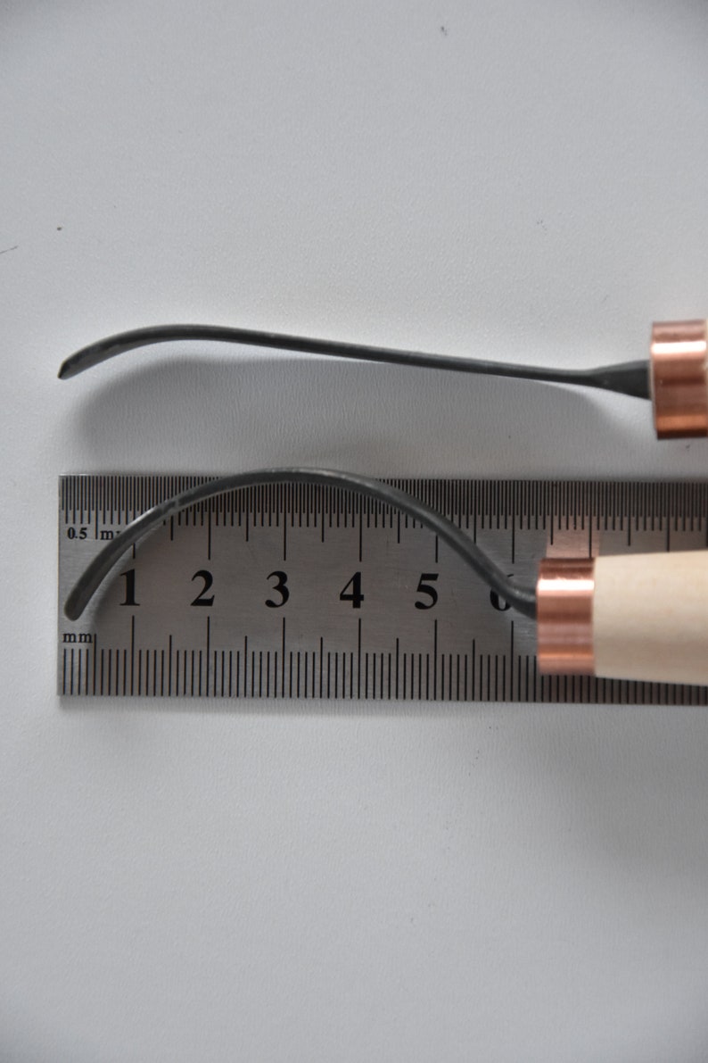 May include: Two wood carving tools with copper accents. The tools are laid out on a ruler with markings in millimetres. The top tool is 6.5 cm long and the bottom tool is 7 cm long.