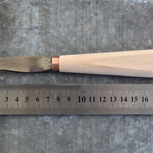 May include: A wooden-handled knife with a copper ferrule and a metal blade. The blade is approximately 20 cm long. The handle has a circular logo with the letter "D" inside.