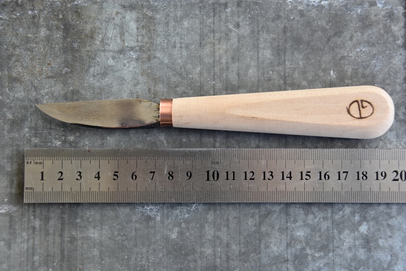 May include: A wooden-handled knife with a copper ferrule and a metal blade. The blade is approximately 20 cm long. The handle has a circular logo with the letter "D" inside.