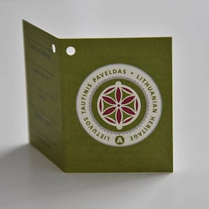 May include: A green tag with a white circle containing a red and white geometric design. The text on the tag reads "Tautinis Paveldass - Lithuanian Heritage - Lietuvos"