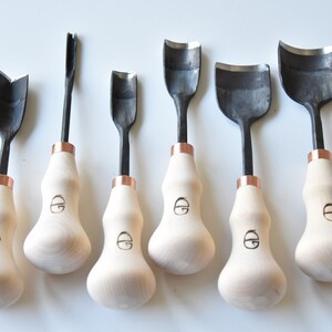 May include: Set of six wood carving tools with black metal blades and wooden handles. The handles are a light wood colour with a copper band at the top. The blades are different shapes, including a V-shape, a U-shape, and a curved shape.