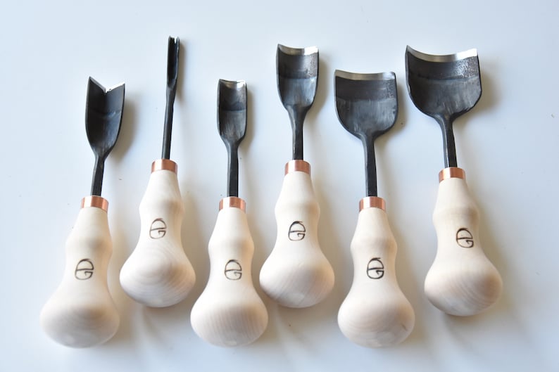 May include: Set of six wood carving tools with black metal blades and wooden handles. The handles are a light wood colour with a copper band at the top. The blades are different shapes, including a V-shape, a U-shape, and a curved shape.