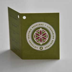 May include: A green tag with a white circle containing a red and white geometric design. The text on the tag reads "Tautinis Paveldas - Lithuanian Heritage - Lietuvos"