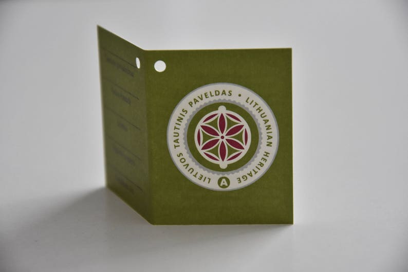 May include: A green tag with a white circle containing a red and white geometric design. The text on the tag reads "Tautinis Paveldas - Lithuanian Heritage - Lietuvos"