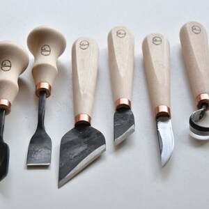 May include: Set of six wood carving tools with wooden handles and copper accents. The tools have different blade shapes for various carving techniques.