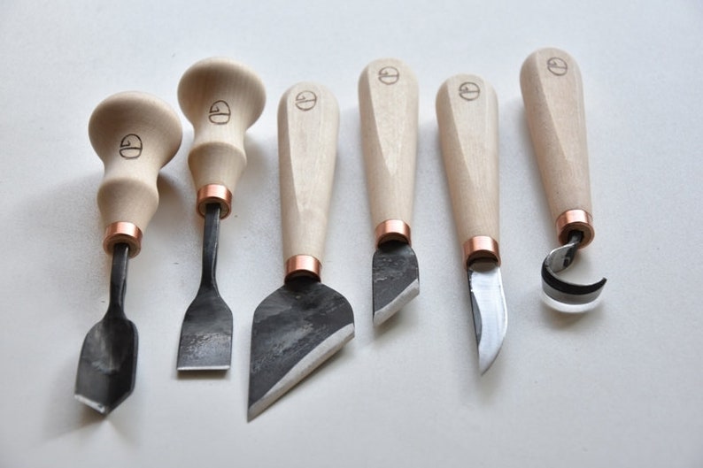 May include: Set of six wood carving tools with wooden handles and copper accents. The tools have different blade shapes for various carving techniques.