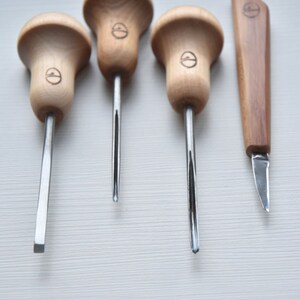 Puede incluir: Cuatro herramientas de talla con mango de madera y cuchillas de metal, y un peque&ntilde;o cuchillo de madera con una cuchilla de metal. Las herramientas tienen un mango en forma de hongo con un logotipo circular.