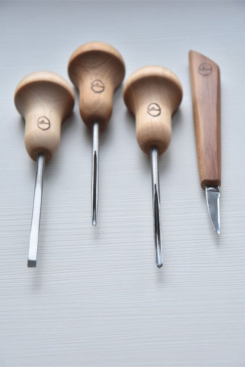 Puede incluir: Cuatro herramientas de talla con mango de madera y cuchillas de metal, y un peque&ntilde;o cuchillo de madera con una cuchilla de metal. Las herramientas tienen un mango en forma de hongo con un logotipo circular.