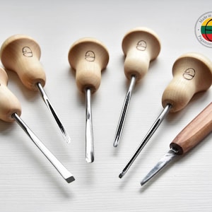 May include: A set of wood carving tools featuring light-coloured wooden handles and stainless steel blades. Each handle has a circular top with a small engraved design. One tool has a rectangular handle. The tools are arranged on a white surface.