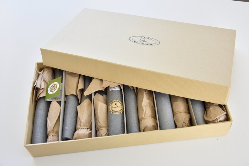 May include: A cardboard box with a lid open, revealing a set of 10 gray cylindrical objects wrapped in brown paper. The objects are arranged in rows within the box. The box is labeled "Handmade" and "Liberty Craft Supplies".