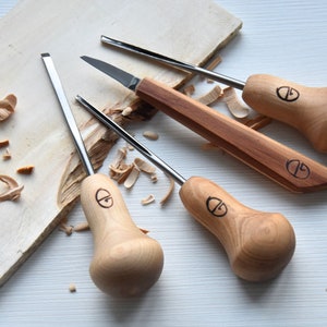 Puede incluir: Conjunto de cuatro herramientas de talla de madera con mangos de madera. Las herramientas tienen un mango en forma de pera con una hoja de metal. Las herramientas est&aacute;n sobre una superficie de madera clara con virutas de madera.