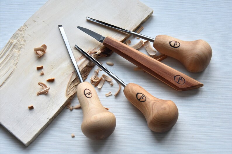 Puede incluir: Conjunto de cuatro herramientas de talla de madera con mangos de madera. Las herramientas tienen un mango en forma de pera con una hoja de metal. Las herramientas est&aacute;n sobre una superficie de madera clara con virutas de madera.