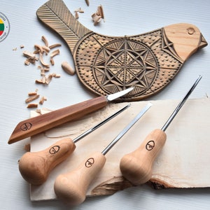 May include: A set of four wood carving tools with wooden handles. The tools have a variety of blade shapes and sizes. The tools are laid out on a piece of wood with wood shavings scattered around. A wooden bird carving is in the background.