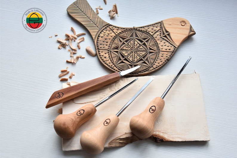 Puede incluir: Un juego de cuatro herramientas de talla de madera con mangos de madera. Las herramientas tienen una variedad de formas y tama&ntilde;os de cuchillas. Las herramientas est&aacute;n dispuestas sobre una pieza de madera con virutas de madera esparcidas alrededor. Una talla de p&aacute;jaro de madera est&aacute; en el fondo.