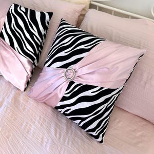 May include: Two square throw pillows featuring a black and white zebra print pattern combined with a soft pink satin fabric. A gathered pink satin band crosses the center of each pillow, secured by a circular silver-toned brooch with clear crystals.