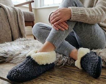 May include: Cozy, dark gray slippers with a cream-colored lining. The slippers are made of a soft, textured material and have a dark blue sole. The image shows the slippers on a wooden floor, suggesting comfort and relaxation.
