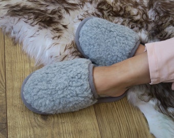 May include: A pair of gray fuzzy slippers with a soft, plush texture. The slippers are worn by a person sitting on a fluffy rug.