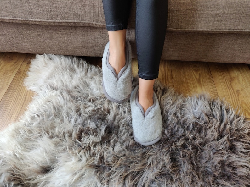 May include: A pair of grey fluffy slippers with a V-shaped opening at the top. The slippers are on a fluffy grey rug.