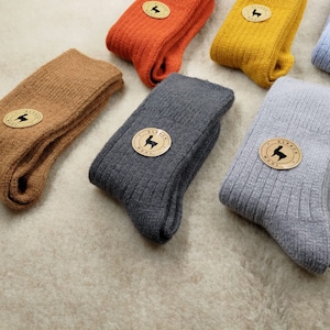 May include: Assortment of ribbed knit socks in various colors, including brown, orange, yellow, gray, and blue. Each pair features a circular label with an alpaca logo. The socks are neatly folded, showcasing their texture and design.