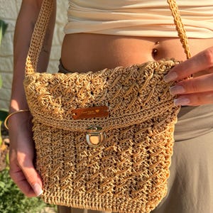 Può includere: Una borsa a tracolla in uncinetto beige con un motivo intrecciato testurizzato, una chiusura a pressione in metallo dorato e una tracolla in uncinetto abbinata. Una piccola etichetta rettangolare in pelle marrone riporta la scritta Hand Made with love.