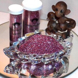Puede incluir: Dos frascos de plástico transparente llenos de purpurina rosa, etiquetados como "Monkey Boy Glitter Tulip Pink". Un plato de vidrio lleno de purpurina rosa se encuentra delante de los frascos, con una pequeña figura de mono de porcelana encima del plato.