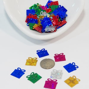 May include: Assortment of colorful, glittery gift box shaped confetti. The confetti is in a white bowl and scattered on a white surface.