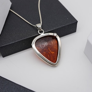 Pode incluir: Um colar com pingente de prata com um pingente em forma de palheta de guitarra. O pingente tem uma aparência marrom marmorizada com um logotipo e é emoldurado em uma moldura prateada. Uma pequena pedra preciosa transparente é colocada na moldura. A corrente do colar também é prateada.