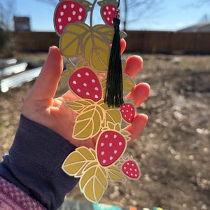 May include: A metal bookmark featuring a strawberry design. The bookmark has red strawberries with white dots, yellow leaves, and a green tassel. The design is held in a hand against a blurred outdoor background.