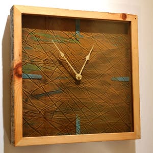 May include: Square wall clock with a natural wood frame. The clock face has a textured brown surface with blue and green accents and gold-colored hands. The clock is approximately 12 inches square.