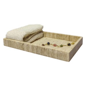 Puede incluir: Una bandeja rectangular hecha de piedra travertina beige claro con una toalla blanca doblada y un brazalete de cadena dorada con cuentas de colores en la parte superior.