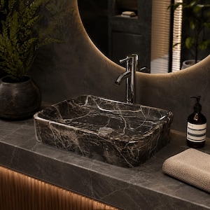 Puede incluir: Un lavabo rectangular de mármol gris oscuro y negro con vetas blancas prominentes. El lavabo presenta un acabado pulido y un desagüe metálico central. Está colocado sobre una encimera de piedra a juego con un grifo de cromo.