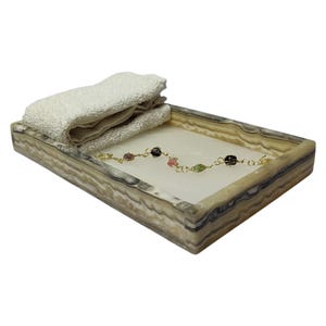 Puede incluir: Una bandeja rectangular hecha de ónix blanco y marrón con una toalla blanca y un brazalete de cadena dorada con piedras de colores y cuentas negras en la parte superior.