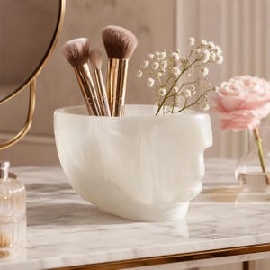 Puede incluir: Un recipiente de resina blanca con efecto mármol en forma de cabeza humana, que contiene tres brochas de maquillaje con mangos dorados metálicos y una rama de pequeñas flores blancas. El recipiente tiene un acabado liso y brillante.
