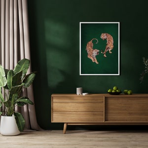 Op de afbeelding: Een ingelijste print van twee tijgers die elkaar aankijken op een groene achtergrond. De tijgers staan in een dynamische pose, met hun staarten om hun lichaam gekruld. De print hangt aan een muur boven een houten dressoir.