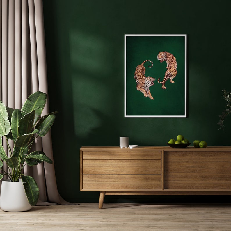 Op de afbeelding: Een ingelijste print van twee tijgers die elkaar aankijken op een groene achtergrond. De tijgers staan in een dynamische pose, met hun staarten om hun lichaam gekruld. De print hangt aan een muur boven een houten dressoir.