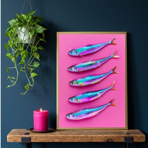 May include: A framed print featuring five iridescent fish arranged vertically on a vibrant pink background. The fish have blue, purple, and green hues. The frame is gold. A pink candle sits on a wooden shelf.