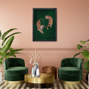 Op de afbeelding: Een ingelijste print van twee tijgers die elkaar aankijken op een groene achtergrond. De tijgers staan in een cirkelvormige opstelling, met hun staarten in elkaar gevlochten. De print hangt aan een roze muur, boven een gouden en bruine salontafel en twee groene fluwelen fauteuils.