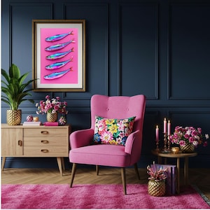 May include: A framed art print featuring five iridescent fish on a pink background. The artwork hangs above a pink armchair with a floral pillow. The room has a dark blue wall, a wooden cabinet, and a pink rug.