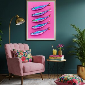 May include: A framed art print featuring five iridescent fish arranged vertically on a vibrant pink background. The fish have blue, purple, and silver hues with orange tail fins. The artwork is displayed in a room with a pink armchair and floral pillow.
