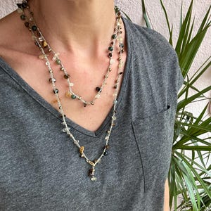 Puede incluir: Un collar de varias capas con pequeñas piedras preciosas de colores y cuentas doradas ensartadas en una cadena plateada. El collar tiene un colgante con un grupo de piedras. La persona lleva una camiseta gris con cuello en V.