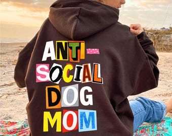 May include: A dark brown hoodie with the words "ANTI SOCIAL DOG MOM" in colorful, cut-out style letters. The hoodie is worn by a person sitting on a beach, with a colorful towel beneath them.