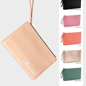 Op de afbeelding: Een blush roze clutch met een witte gepersonaliseerde inscriptie die "Shaema" luidt. De tas heeft een ritssluiting bovenaan en een polsbandje.