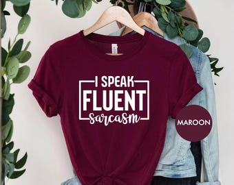 May include: Maroon t-shirt with the text "I Speak Fluent Sarcasm" in a white rectangular box. The shirt is hanging on a wooden hanger. The word "Maroon" is in a circle on the right side of the shirt.